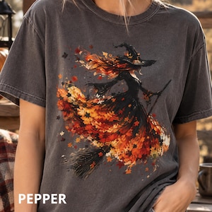 May include: A charcoal gray short-sleeve t-shirt featuring a graphic print of a witch in a pointed hat riding a broomstick. The witch's dress and hair are composed of vibrant orange, red, and yellow autumn leaves. The word PEPPER is printed in white.