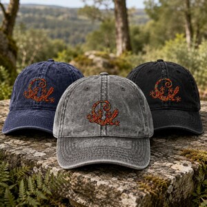 May include: Three baseball caps in navy blue, charcoal grey, and black, each featuring a distressed fabric finish. Centred on the front of each cap is an embroidered design of a stylised orange and red lizard with intricate floral patterns.