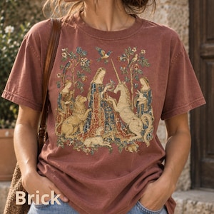 May include: A brick-coloured short-sleeve t-shirt featuring a detailed graphic print inspired by medieval tapestry art. The design depicts a central figure surrounded by a lion, a unicorn, and other animals amidst floral motifs and trees.