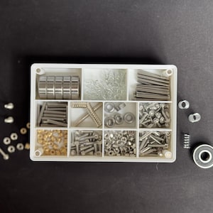 May include: A white compartmented box containing an assortment of small metal hardware components. The box includes bearings, pins, screws, nuts, and other fasteners. A single bearing and other hardware pieces are outside the box on a black surface.
