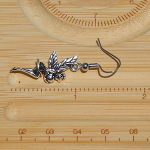 May include: A silver-toned metal earring featuring a winged figure in a reclining pose, attached to a curved ear wire. The earring is positioned against a wooden ruler, showing the figure measures approximately 3.8 cm in length.