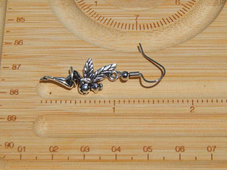 May include: A silver-toned metal earring featuring a winged figure in a reclining pose, attached to a curved ear wire. The earring is positioned against a wooden ruler, showing the figure measures approximately 3.8 cm in length.