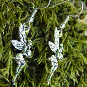 May include: A pair of silver-toned earrings featuring a detailed fairy charm with outstretched wings. Each earring is suspended from a simple silver-toned hook wire, showcasing a polished metallic finish and intricate wing patterns on the fairy figure.