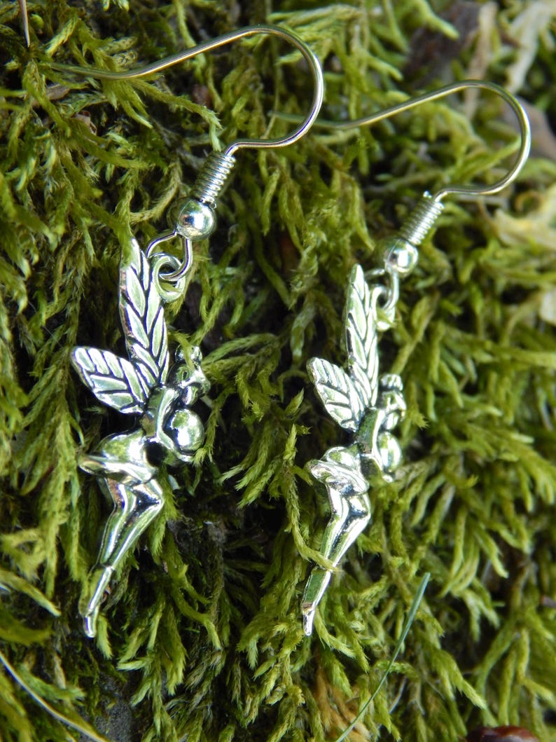 May include: A pair of silver-toned earrings featuring a detailed fairy charm with outstretched wings. Each earring is suspended from a simple silver-toned hook wire, showcasing a polished metallic finish and intricate wing patterns on the fairy figure.