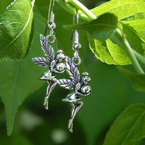 May include: A pair of silver-toned metal earrings featuring a fairy design. Each earring depicts a small figure with wings, holding a spherical object, suspended from a standard hook-style ear wire. The metal has a polished, reflective finish.