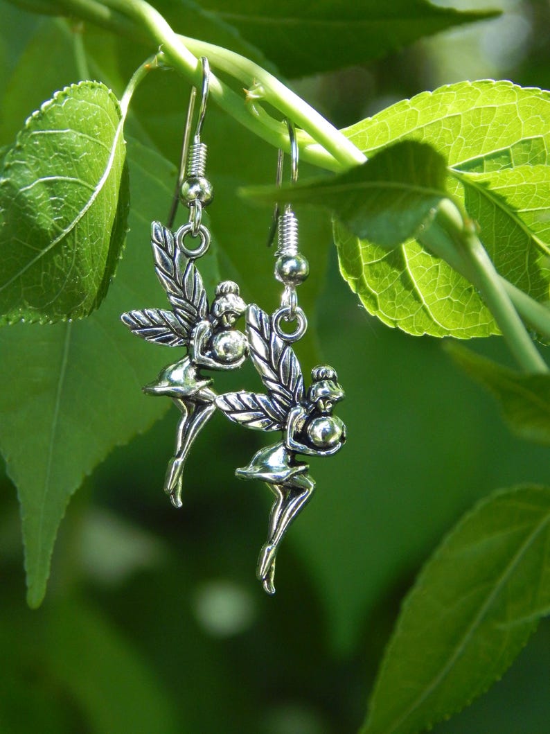 May include: A pair of silver-toned metal earrings featuring a fairy design. Each earring depicts a small figure with wings, holding a spherical object, suspended from a standard hook-style ear wire. The metal has a polished, reflective finish.