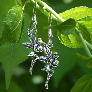 May include: A pair of silver-toned drop earrings featuring a fairy charm design. Each earring depicts a small figure with wings, holding a spherical object, suspended from a standard metal hook wire. The charms have a polished metallic finish.