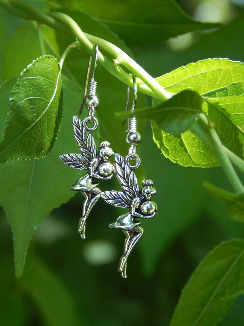May include: A pair of silver-toned drop earrings featuring a fairy charm design. Each earring depicts a small figure with wings, holding a spherical object, suspended from a standard metal hook wire. The charms have a polished metallic finish.