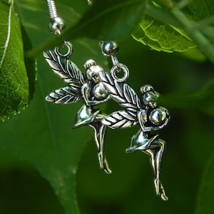 May include: A pair of silver-toned metal earrings featuring a detailed fairy figure with wings. Each earring is suspended from a standard hook wire, showcasing a polished finish that reflects light, depicting the fairy in a seated pose.