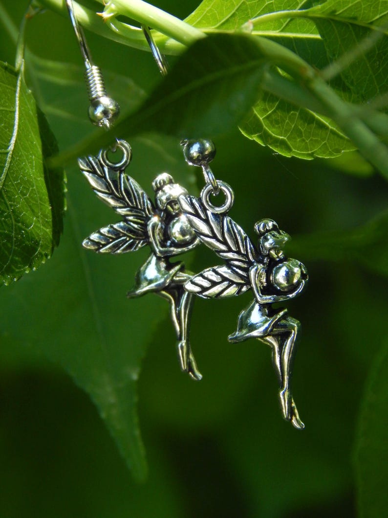 May include: A pair of silver-toned metal earrings featuring a detailed fairy figure with wings. Each earring is suspended from a standard hook wire, showcasing a polished finish that reflects light, depicting the fairy in a seated pose.