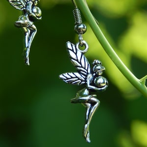 May include: A pair of silver-toned drop earrings featuring a detailed fairy figure holding a small sphere. Each earring has a hook-style fastening and displays a winged character with defined features, suspended from a thin metal wire loop.