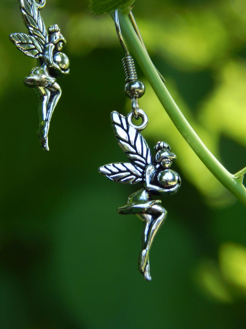 May include: A pair of silver-toned drop earrings featuring a detailed fairy figure holding a small sphere. Each earring has a hook-style fastening and displays a winged character with defined features, suspended from a thin metal wire loop.