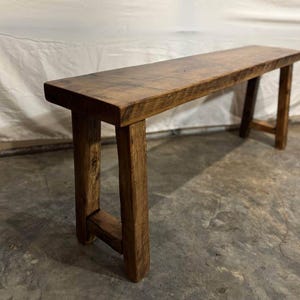 May include: A rustic, rectangular wooden bench with a natural, aged finish. The bench has a long, flat top supported by four sturdy, angled legs. The wood grain is visible, adding to its charm. The bench is approximately 60 inches long.