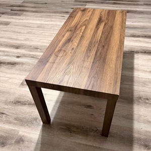 Op de afbeelding: Een rechthoekige houten salontafel met een rijke bruine afwerking. De tafel heeft een eenvoudig, modern ontwerp met vier stevige poten. De houtnerf is zichtbaar en voegt textuur en warmte toe. De tafel is ongeveer 122 cm lang en 51 cm breed.
