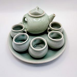 May include: A celadon green ceramic tea set consisting of a teapot with a lid and five small, rounded tea cups arranged on a matching circular tray. The pieces feature a smooth, glossy glaze with a subtle, uniform light green color throughout.