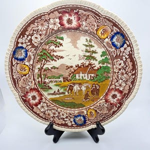 May include: A round ceramic plate with a scalloped edge featuring a central landscape illustration of a house, a river with a boat, and figures with horses. The border is decorated with brown floral patterns and colorful blue, red, and yellow flowers.
