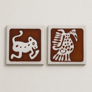May include: Two square ceramic tiles featuring a white graphic design on a deep brown background. The left tile depicts a stylized four-legged animal with a curved tail, while the right tile shows a bird with detailed wing and body patterns.