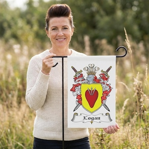 Puede incluir: Una bandera de jardín blanca con un escudo familiar. El escudo presenta un escudo amarillo con un corazón rojo, espadas cruzadas, un yelmo y el nombre "Logan" en una pancarta. La bandera se sostiene con un soporte de metal negro.