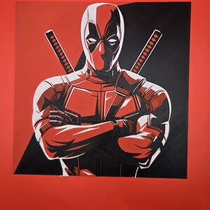 May include: A square graphic print featuring a stylised illustration of the character Deadpool with arms crossed. The character wears a red and black suit and mask, with two katana sword hilts visible behind the shoulders against a split background.