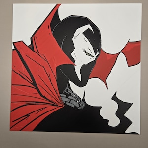 May include: A square graphic art print featuring a stylised illustration of a comic book character with a black mask, white eye markings, and a large, flowing red cape. The character is depicted in profile against a plain white background.
