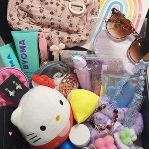 May include: A pink corduroy backpack with a floral pattern, a Byoma lotion tube, brown sunglasses, a Hello Kitty plush, a yellow cheese-shaped sponge, butterfly earrings, a beaded bracelet, a purple hair tool, and various small accessories in a box.