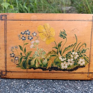 May include: A rectangular wooden box with a light brown finish featuring a hand-painted floral design. The artwork displays a large yellow pansy, small blue forget-me-nots, and white blossoms with green leaves. A thin black border frames the entire top.