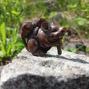 May include: A dark brown, hand-carved wooden figurine of a mouse. The mouse is depicted in a seated position, carrying a small, rounded bundle on its back and holding a thin, cylindrical object with its front paws, featuring detailed textures.