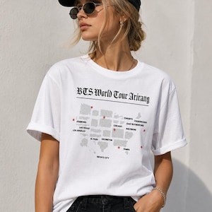 Könnte beinhalten: Ein weißes kurzärmeliges T-Shirt mit dem Text BTS World Tour Arirang über einer grauen Karte von Nordamerika. Die Karte enthält rote Sternsymbole und Städtenamen wie Stanford, Las Vegas, Los Angeles, El Paso, Arlington, Chicago und Toronto.