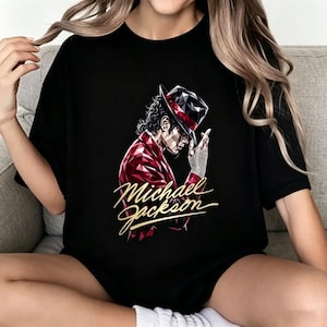 以下が含まれることがあります： マイケル・ジャクソンのイラストが描かれた黒いTシャツ。赤と白の幾何学模様のジャケットと帽子、銀色の手袋をしたジャクソンが描かれています。「Michael Jackson」の文字は金色の筆記体で書かれています。