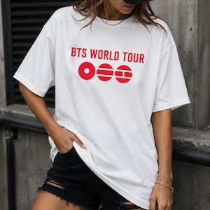 May include: White t-shirt with the text "BTS WORLD TOUR" in red, along with three red circular graphic elements. The shirt is paired with black denim shorts. The person is wearing a black cap.
