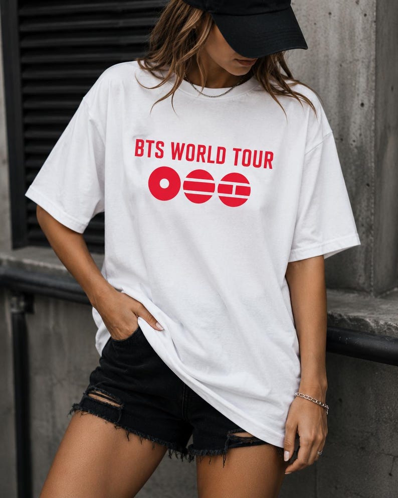 May include: White t-shirt with the text "BTS WORLD TOUR" in red, along with three red circular graphic elements. The shirt is paired with black denim shorts. The person is wearing a black cap.