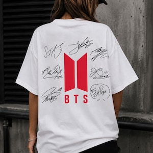 May include: White t-shirt featuring a red and white BTS logo with signatures. The shirt has a crew neck and short sleeves. The BTS logo is centered on the back of the shirt.