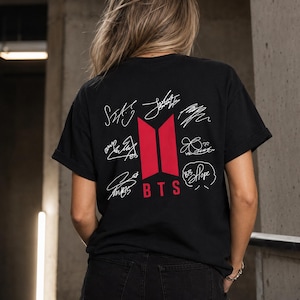 May include: Black t-shirt featuring the BTS logo in red and white signatures. The shirt is worn by a person with blonde hair and black denim shorts. The shirt's design includes the band's logo and the word "BTS".