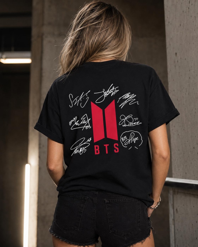 May include: Black t-shirt featuring the BTS logo in red and white signatures. The shirt is worn by a person with blonde hair and black denim shorts. The shirt's design includes the band's logo and the word "BTS".