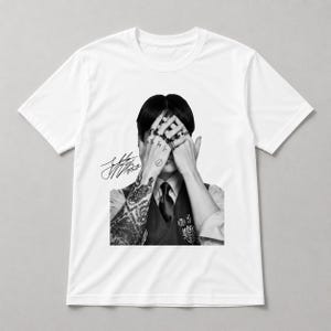 Puede incluir: Camiseta blanca con una imagen en blanco y negro de una persona con tatuajes y anillos, cubriéndose los ojos. La camiseta también incluye una firma y texto.