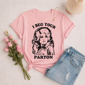 Könnte beinhalten: Ein hellrosa T-Shirt mit kurzen Ärmeln und einer schwarzen grafischen Illustration einer Frau mit voluminösem Haar sowie dem Text I BEG YOUR PARTON, der in fetter, geschwungener Schrift über und unter dem Porträt gedruckt ist.