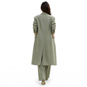 Pu&ograve; includere: Un cappotto lungo verde salvia e pantaloni abbinati. Il cappotto ha un design classico con revers a lancia, polsini con bottoni e tasche. I pantaloni sono a gamba larga. L'outfit &egrave; indossato con tacchi alti verdi.