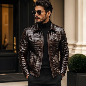 Puede incluir: Chaqueta de cuero marrón con textura de cocodrilo. La chaqueta tiene cuello, dos bolsillos en el pecho y cierre de cremallera. La persona lleva un cuello alto negro y gafas de sol.