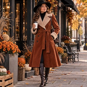 May include: A long, rust-colored corduroy trench coat featuring a wide, cream-colored faux shearling lapel and matching cuffs. The coat has a double-breasted button closure, a tie-waist belt, and is paired with a black wide-brimmed hat and boots.