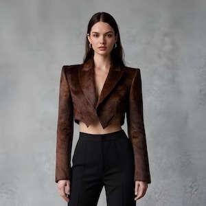 May include: A cropped brown blazer with a notched lapel and long sleeves. The blazer is paired with black high-waisted pants. The outfit is set against a gray background. The blazer's texture appears to be a soft, possibly fur-like material.