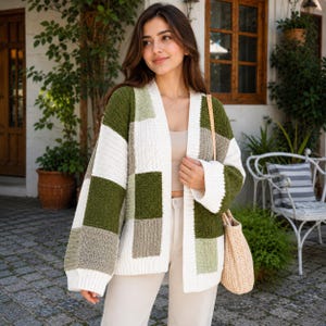 May include: A long-sleeve open-front cardigan featuring a patchwork design of large squares in olive green, cream, and light sage. The knit garment has a ribbed texture, wide sleeves, and a straight hem, paired with a beige woven shoulder bag.