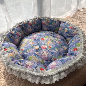 Puede incluir: Una cama redonda para mascotas con un estampado floral en tonos azules, morados y rojos. La cama tiene un ribete de encaje blanco y un interior suave y acolchado, que proporciona un espacio acogedor para una mascota pequeña.