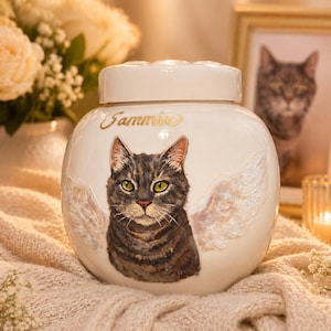 Puede incluir: Urna para mascotas de cerámica color crema con un retrato pintado de un gato atigrado y alas de ángel. La urna tiene tapa y el nombre "Sammie" en escritura dorada. Un retrato enmarcado de un gato está en el fondo.