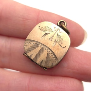 May include: A small, antique gold-colored locket with an engraved floral design. The square locket has a hinged opening and a small loop for attaching to a chain. The engraved details include stylized leaves and lines.