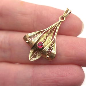 May include: A gold-toned pendant with a floral design. The pendant features three stylized, openwork petals, each with a delicate, curved pattern. A small, round, red gemstone is set in the center. The pendant is approximately 1.5 inches long.