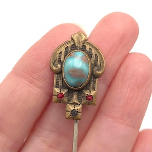 May include: Antique hat pin with a brass setting. The pin features an oval, turquoise-colored stone with brown marbling. The brass setting has an Art Nouveau design with red and black accents. The pin is held by a hand.
