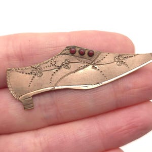 May include: A vintage gold-toned shoe-shaped brooch. The brooch features intricate etched floral designs and three small red stones near the top. The shoe has a pointed toe and a small heel, showcasing a classic, antique aesthetic.