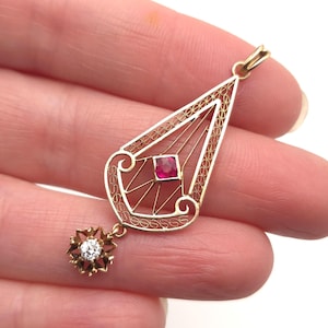 May include: A gold pendant with a filigree design. The pendant features a red square gemstone and a small, flower-shaped charm with a clear gemstone. The pendant is held in a hand.