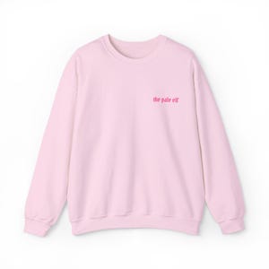 Op de afbeelding: Een lichtroze sweatshirt met lange mouwen. De tekst "the pale elf" is in een donkerdere roze kleur op de bovenborst gedrukt. De sweatshirt is gemaakt van een zacht materiaal.