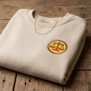 Peut inclure: Un sweat-shirt à col rond beige avec un écusson brodé circulaire sur la poitrine. L'écusson présente un symbole de balance de justice rose vif sur fond jaune, entouré d'une bordure marron. Une fine chaîne dorée repose au niveau de l'encolure.